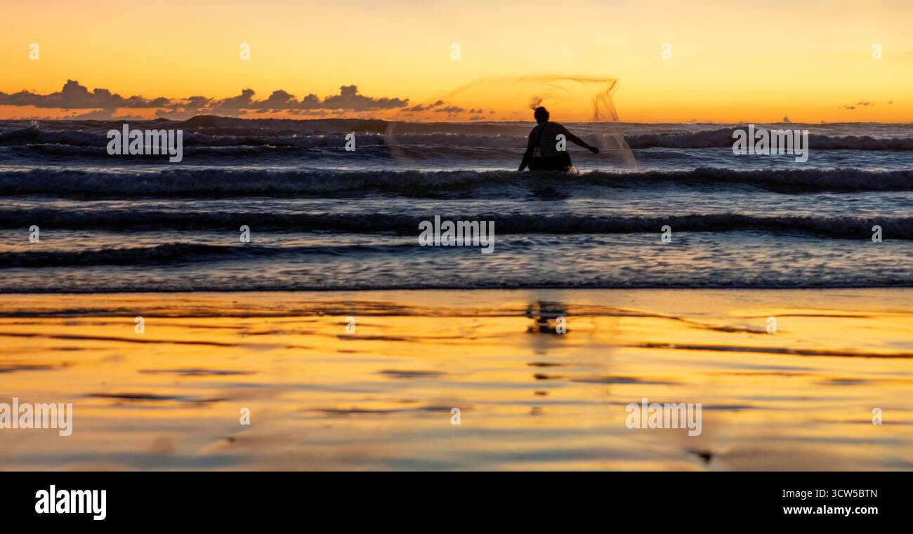 Balinesische Fischer, die bei Sonnenuntergang am Seminyak Beach, Bali, Indonesien, ein Netz ins Surfen werfen Stockfoto
