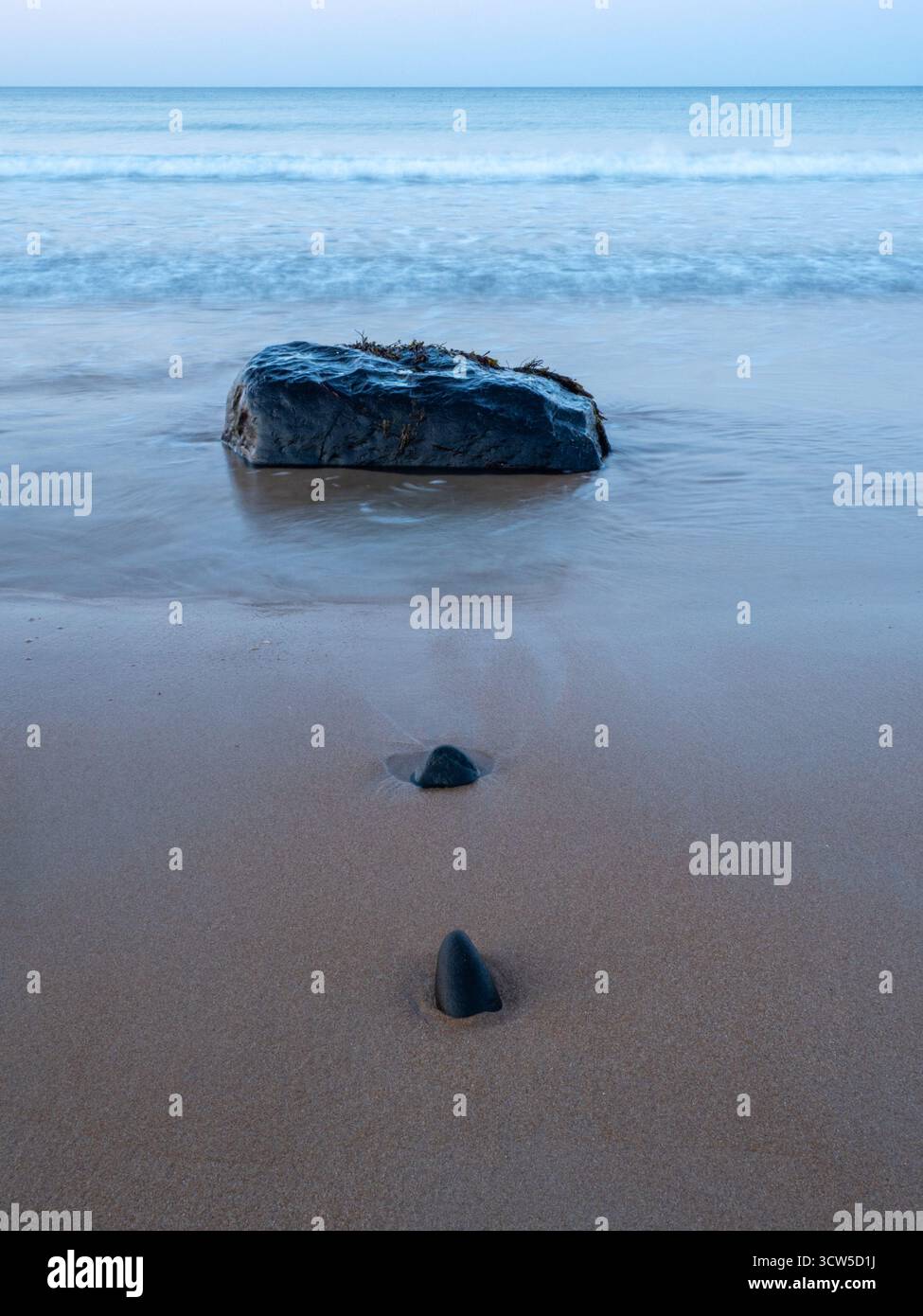 Einzelner großer Felsen und zwei kleinere Steine am Strand mit sanften Wellen und ruhigem Meer Stockfoto