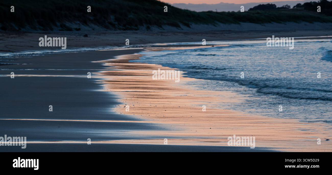Nasser Sand reflektiert sanfte Sonnenaufgangsfarben entlang der ruhigen Strandküste Stockfoto