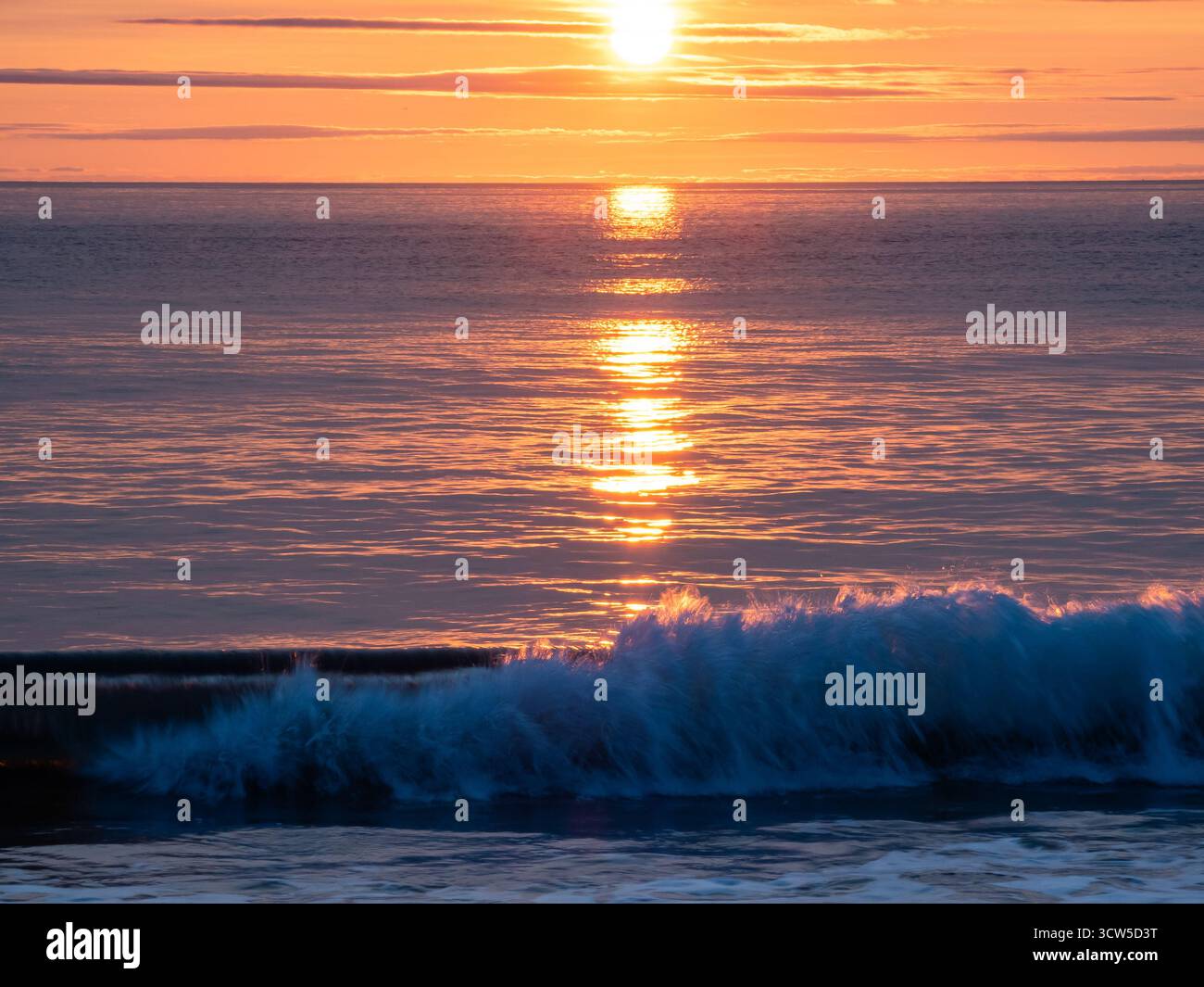 Eine brechende Welle unter dem goldenen Sonnenaufgang, die sich über das ruhige Meer reflektiert Stockfoto