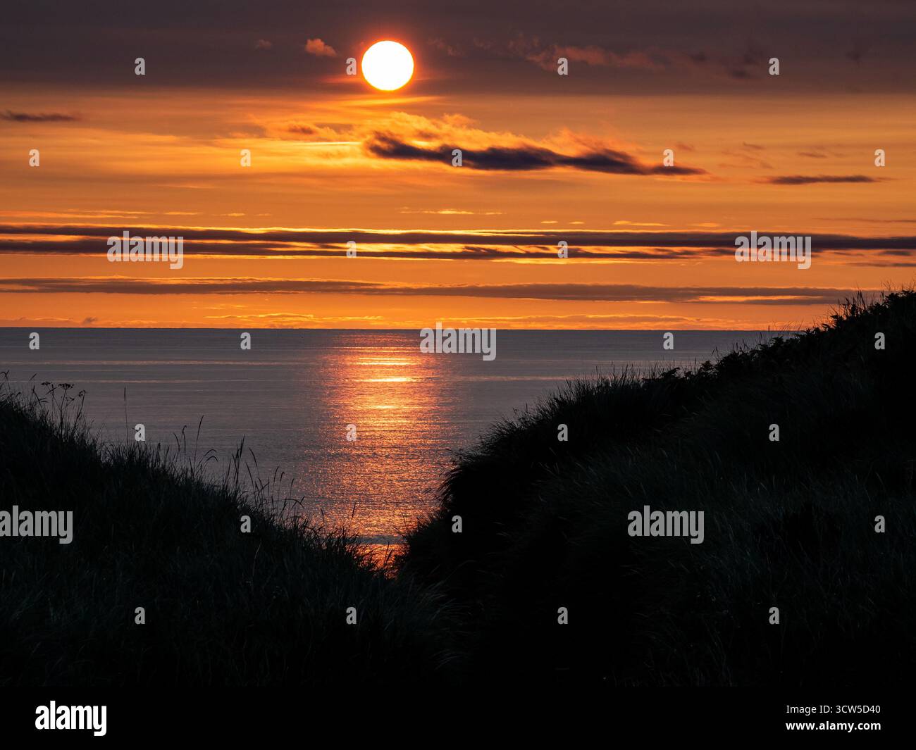 Sonnenaufgang über ruhigem Meer zwischen grasbewachsenen Dünen mit leuchtendem orangefarbenem Himmel Stockfoto