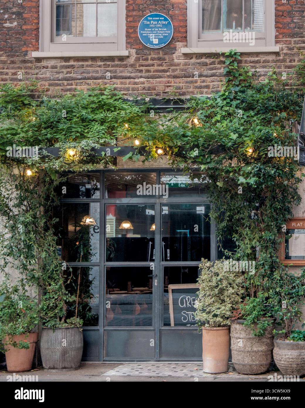 Bezaubernder, mit Efeu bedeckter Eingang des Giaconda Cafe in der Tin Pan Alley, London, mit warmen Lichtern, Grün und einer alten Backsteinfassade. Stockfoto