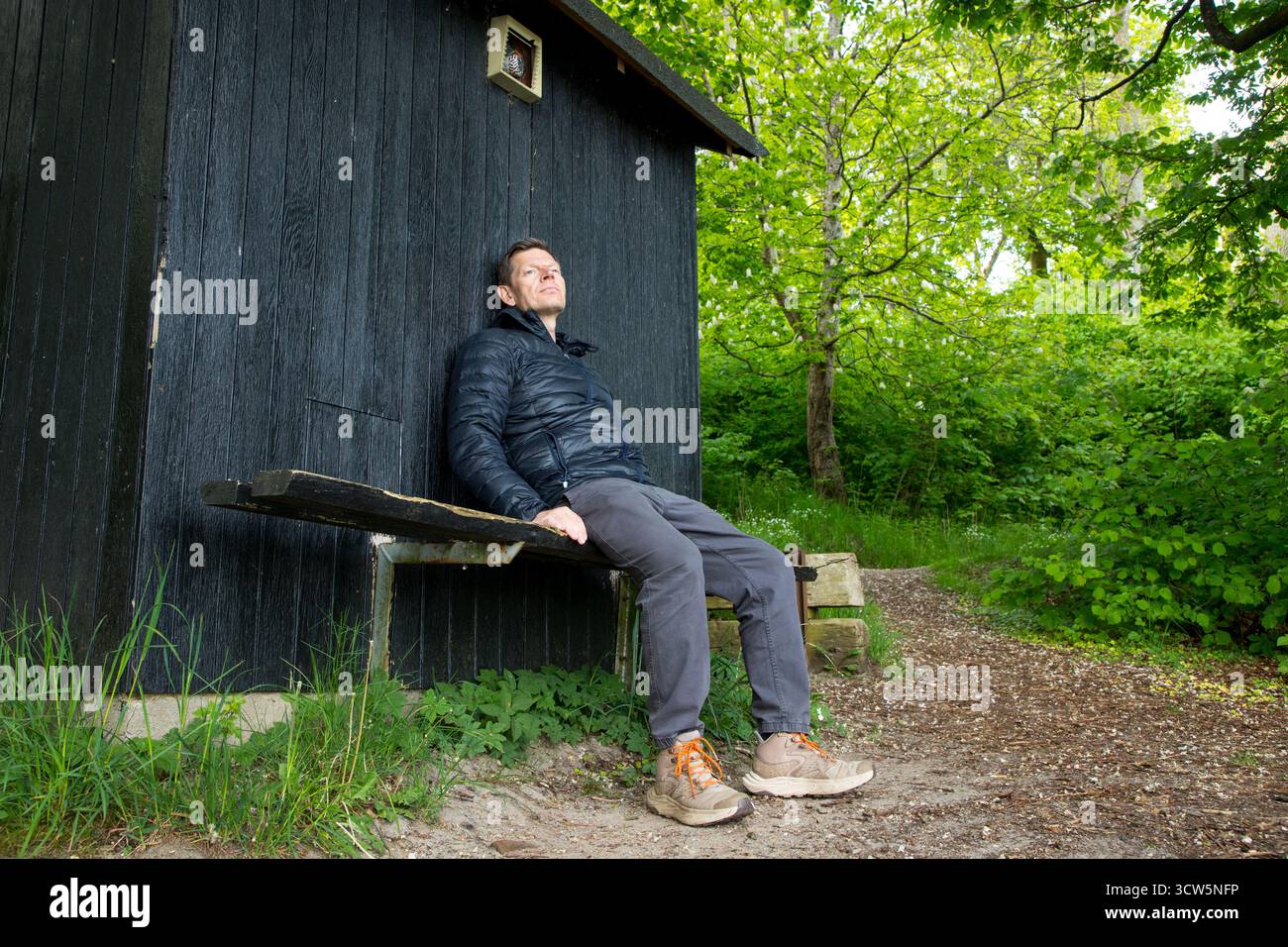 Mann im friedlichen Moment auf Bank im Wald genießt die Natur, Mann lehnt sich an Holzschuppen und glücklich im Naturmoment Stockfoto