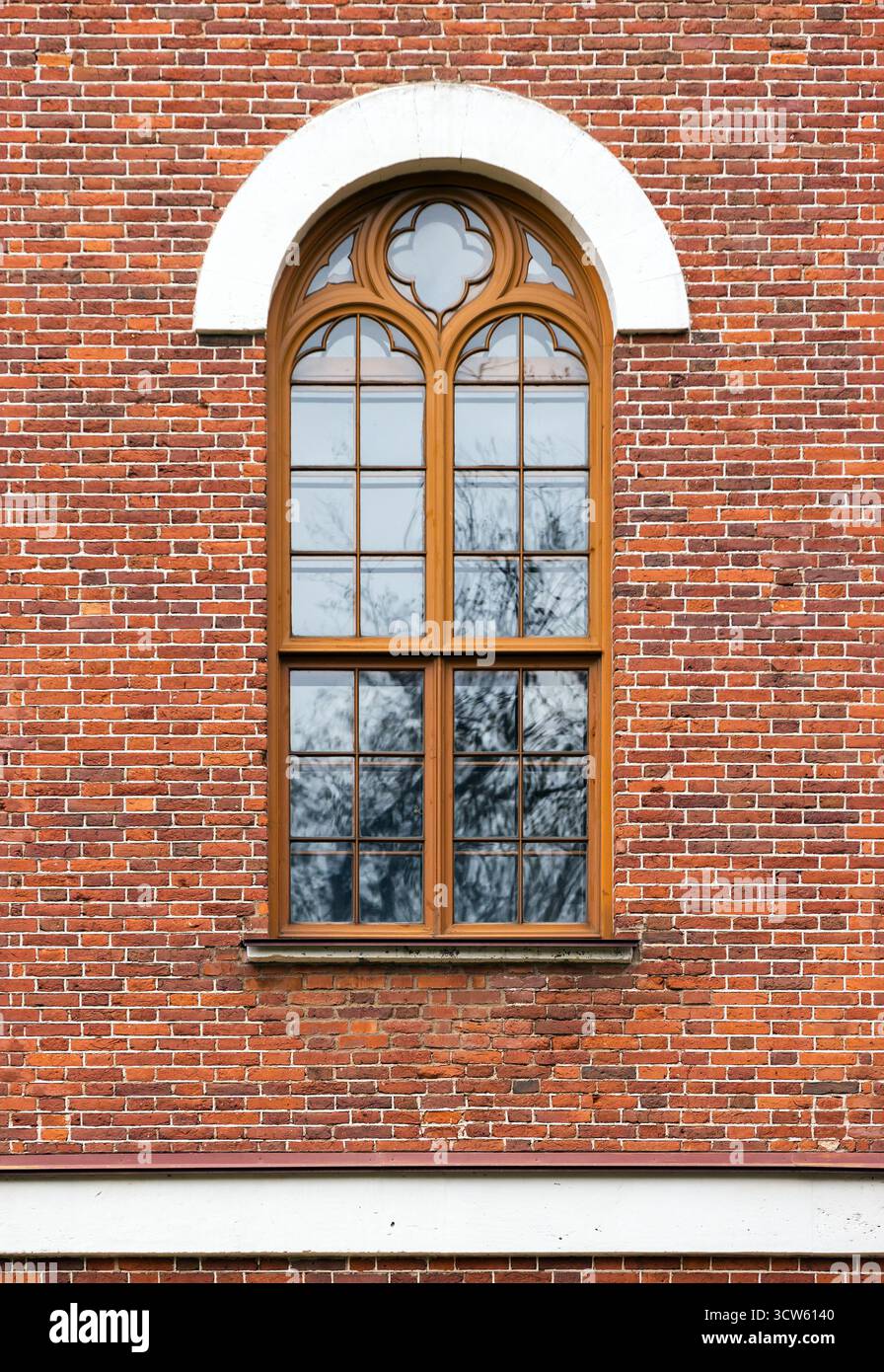Ein vertikales Nahfoto eines verwitterten roten Backsteins mit einem Holzfenster mit gotischen Details und weißem Bogen. Vorderansicht, Hinteransicht Stockfoto