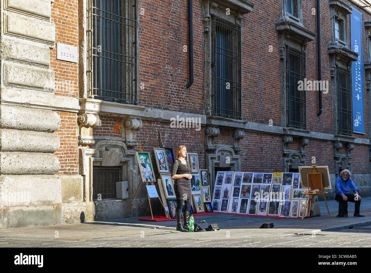 Ein Opernsänger, der in der Via Brera vor der Brera Art Gallery auftritt, mit einem Straßenmaler, der seine Werke im Hintergrund verkauft, in Mailand, Italien Stockfoto