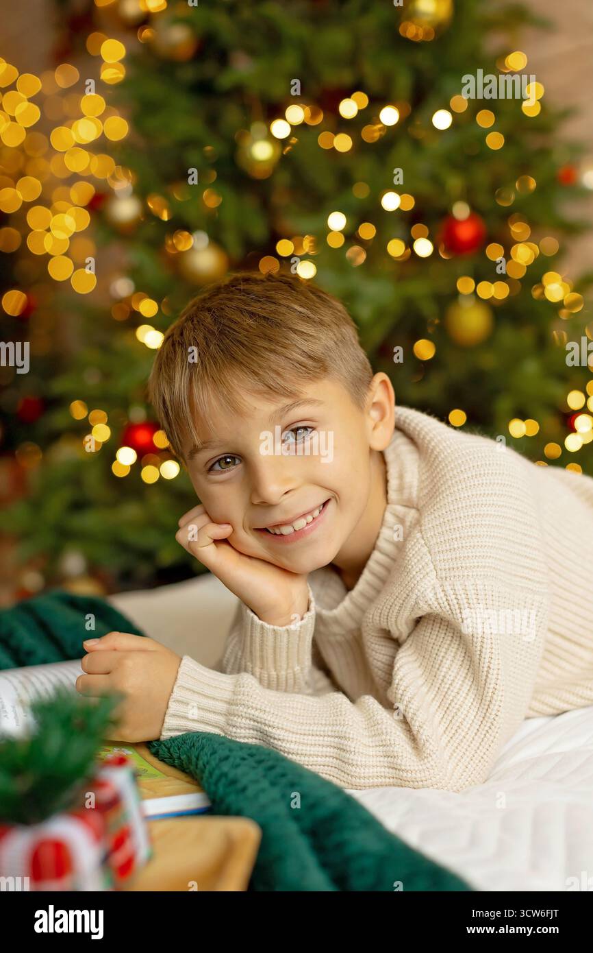 Niedlicher Vorschuljunge, der vor dem Kamin sitzt, weihnachtsgeschenk hält und ein Buch liest, Freude und Glück zu Hause Stockfoto