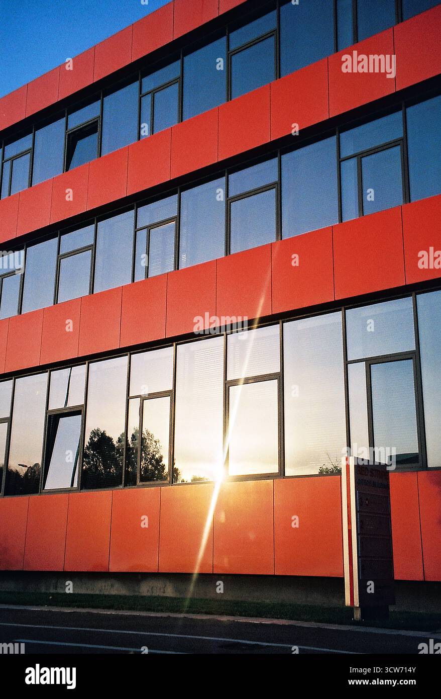 Fassade eines modernen Bürogebäudes mit roten Paneelen und blau reflektierenden Fenstern. Himmel und Sonne spiegeln sich im Glas. Scharfe, saubere Geometrie. Stockfoto