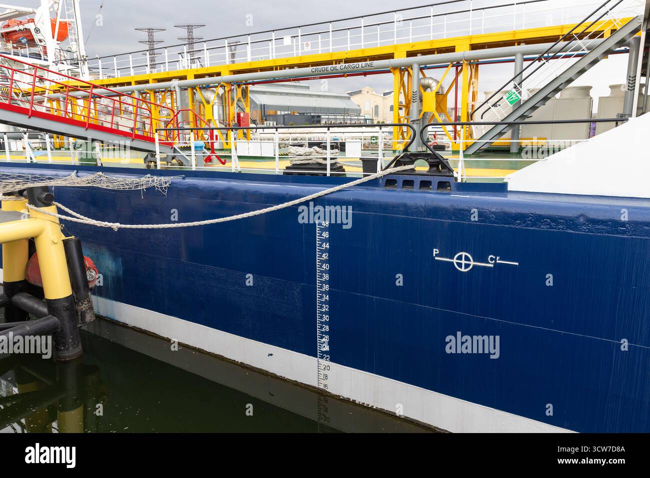 Ein geschäftiges Hafengelände zeigt ein großes blaues Tankschiff, das am Dock mit gelben Rohren und Industrieplattformen vor Anker liegt, die eine dynamische Logistik schaffen Stockfoto