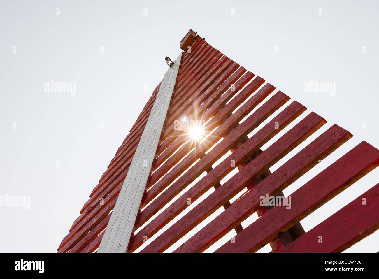 Roter Leuchtturm mit vertikalem weißem Streifen steht unter hellem Himmel, die Sonne leuchtet durch das Holzgerüst, Navigationsausrüstung der Marine Stockfoto