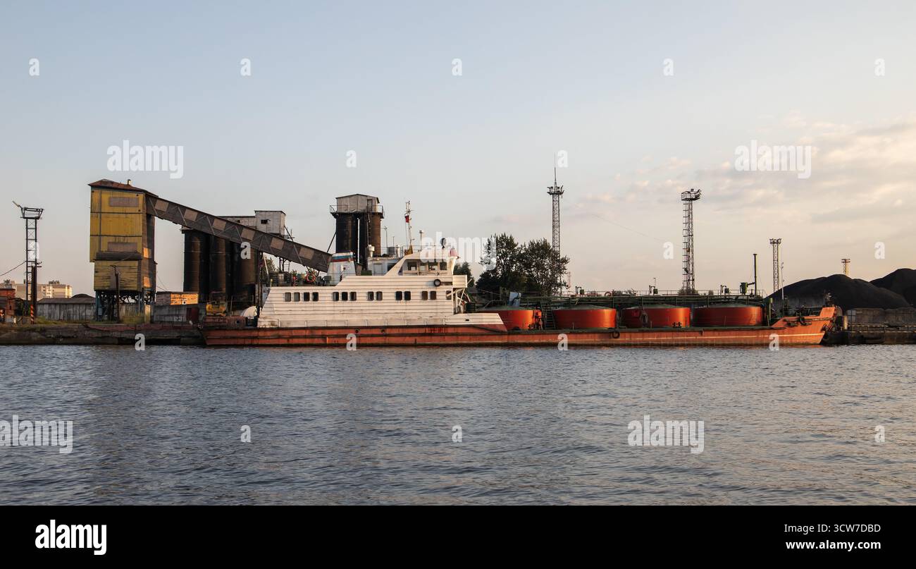 Ein altes Tankschiff sitzt an einem geschäftigen Industriehafen mit großen Förderanlagen, Silos und Kränen entlang des Kais. Die Szene vermittelt Transport, Verladung und m Stockfoto