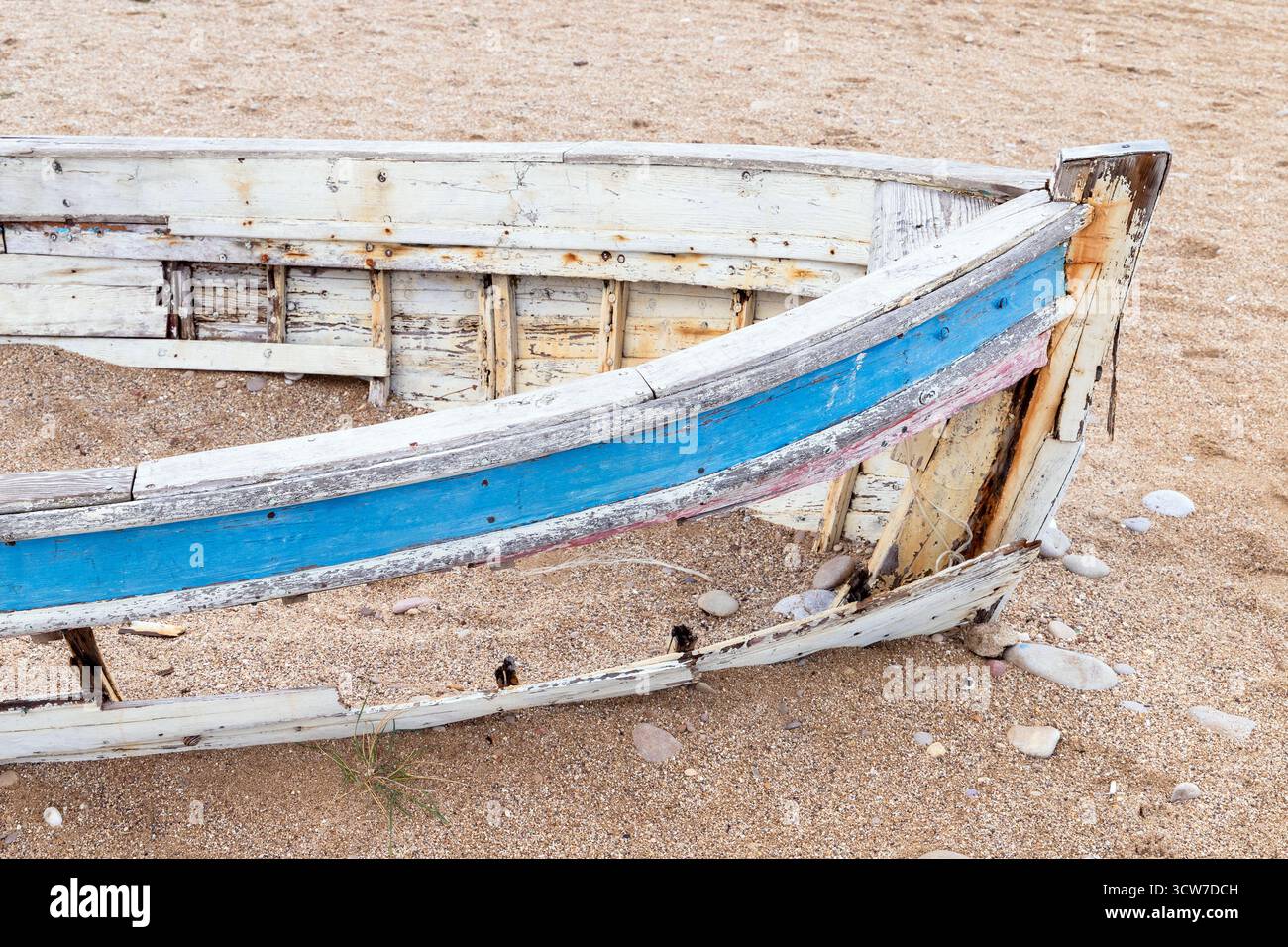 Ein altes, wettergequältes Boot liegt an einem Sandstrand, dessen blauer Streifen sich abblättert. Rostige Nägel, raues Holz und verstreute Steine erinnern an Nostalgie, Solit Stockfoto