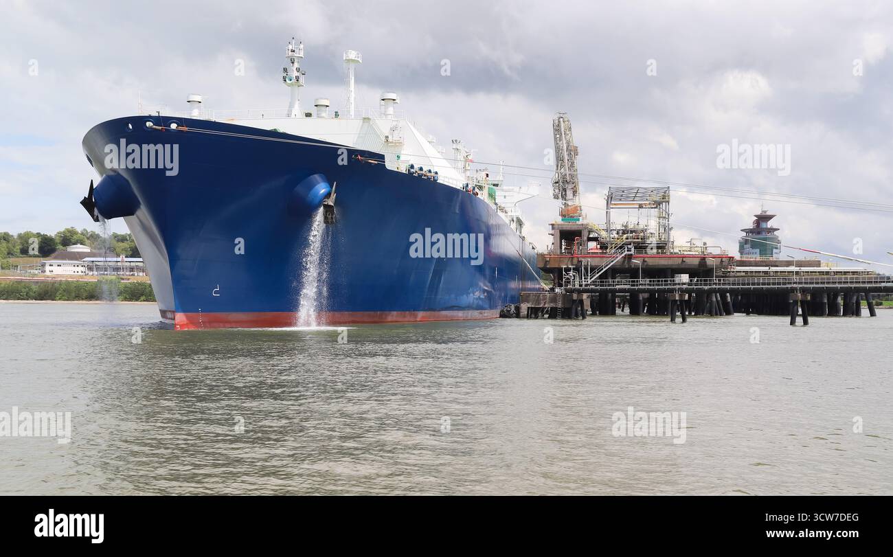 Ein großes Tankschiff mit blauem Rumpf liegt am Ölsteg vor Anker und ist mit einer angedockten Plattform verbunden. Geschäftige Hafenanlage mit Kränen, Wasser und einer Wolke Stockfoto