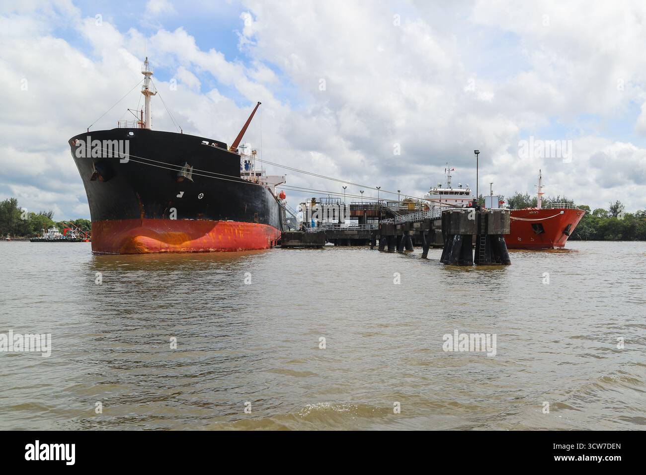 Ein großes schwarzes Tankschiff mit rotem Rumpf liegt an einem verwitterten Ölsteg vor Anker und ist mit einer angedockten Plattform verbunden. Geschäftige Hafenanlage mit Kränen, Wasser Stockfoto