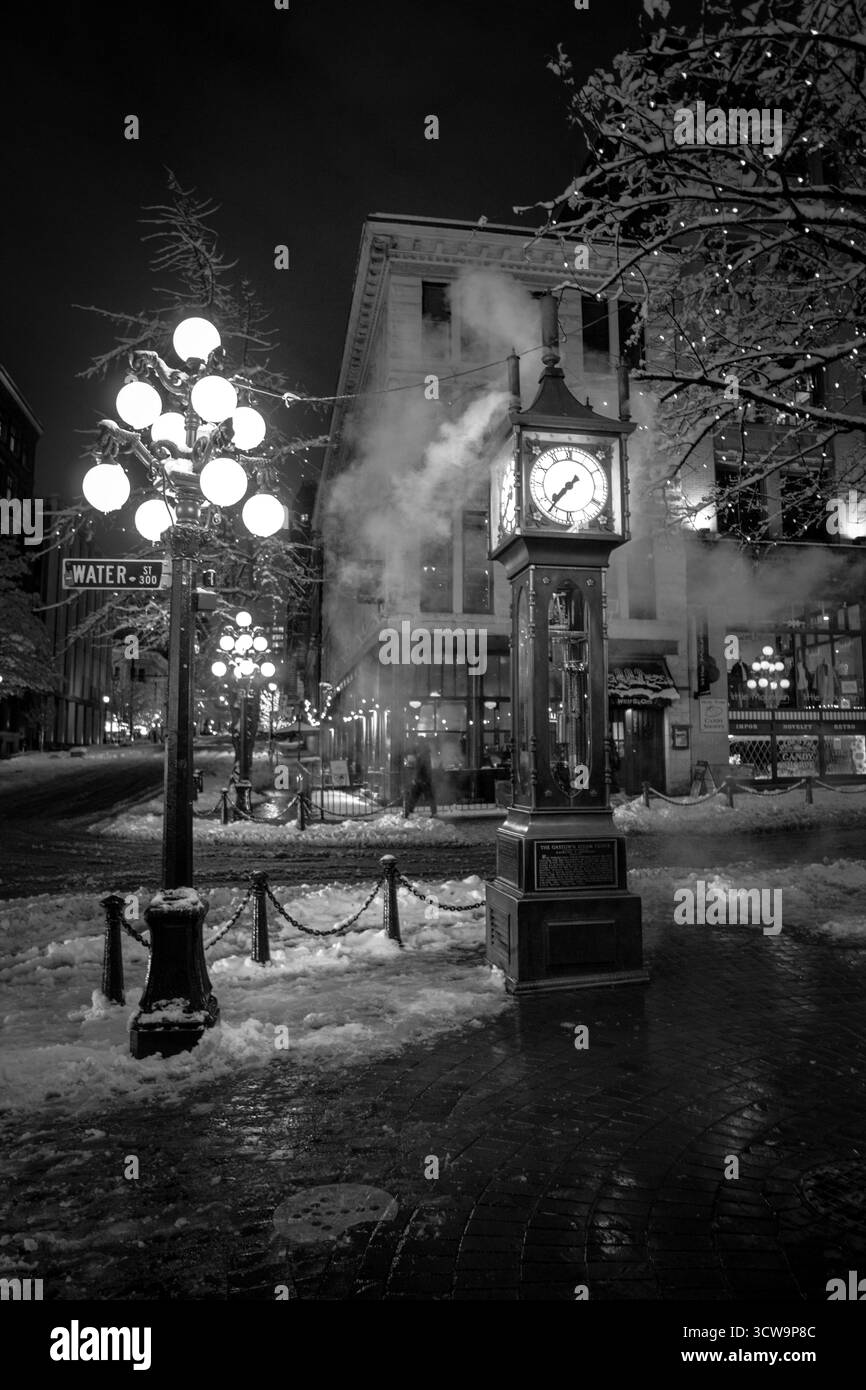 Schnee bedeckt den Boden, während eine Dampfuhr hoch steht. Straßenlaternen beleuchten die Gegend und schaffen eine magische Atmosphäre. Die Leute laufen in der ruhigen Stadt vorbei Stockfoto