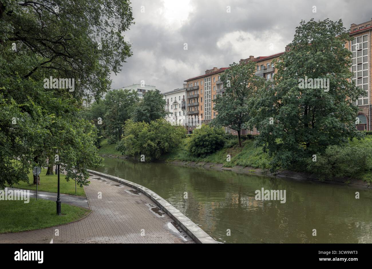 Ein ruhiger Blick auf den Fluss im Central Park, Minsk, mit einem gepflasterten Fußweg, üppigen grünen Bäumen und mehrstöckigen Wohnhäusern am Wasser Stockfoto
