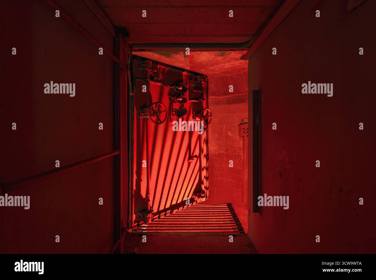 Eine massive verstärkte Sprengkammertür in einer Industrieanlage, teilweise offen und von dramatischem rotem Licht beleuchtet. Stockfoto