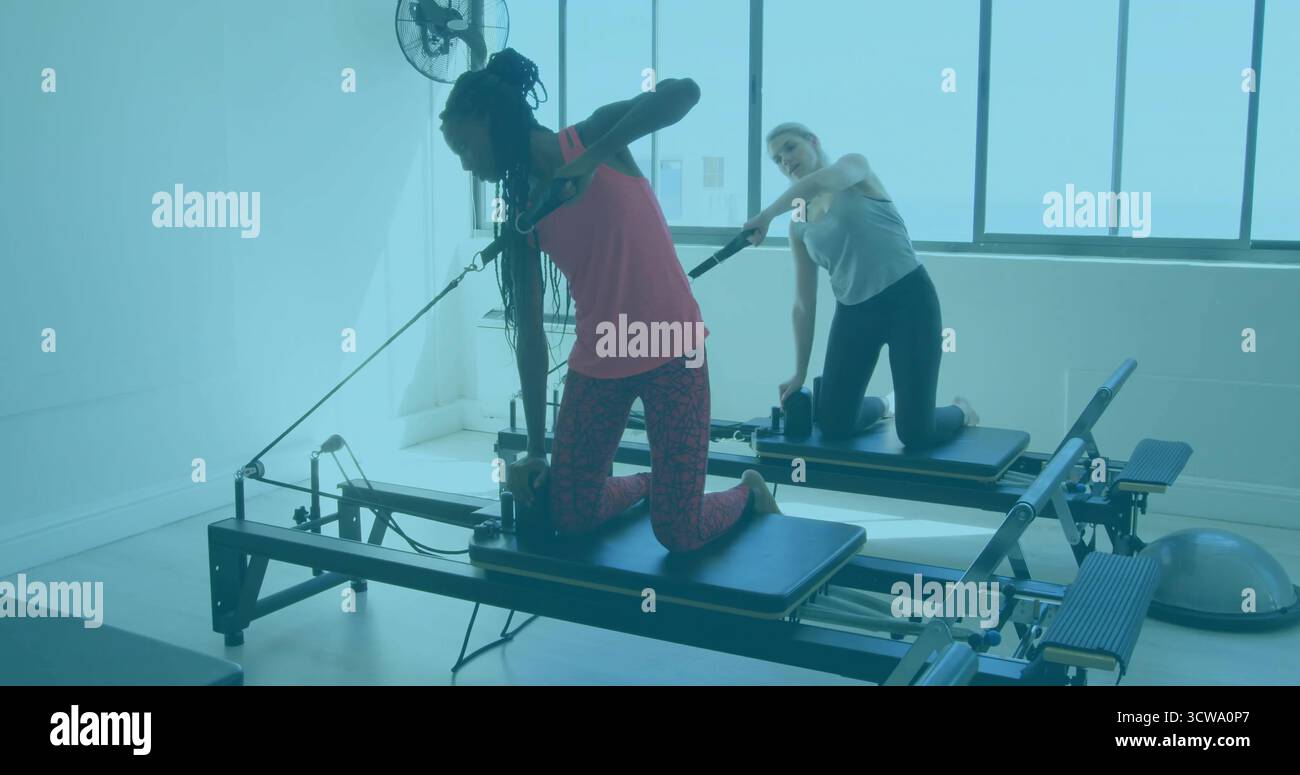 Zwei Frauen in Sportbekleidung knieten bei Sonnenlicht im Pilates-Studio auf Reformern Stockfoto