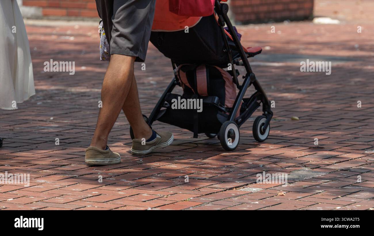Ein Mann in grauen Shorts und legeren Schuhen läuft im Licht, das von umliegenden Bäumen auf die Parkfläche projiziert wird, mit dem Kinderwagen auf rotem Pflaster. Stockfoto