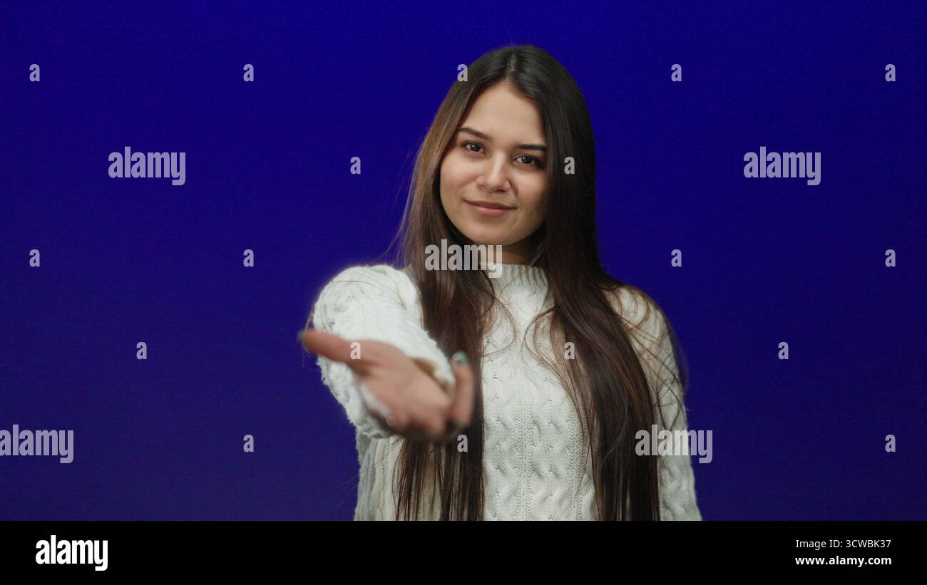 Frau, die über blauem isoliertem Hintergrund zur Kamera greift, trägt einen Pullover mit langen Haaren und einladendem Ausdruck Stockfoto