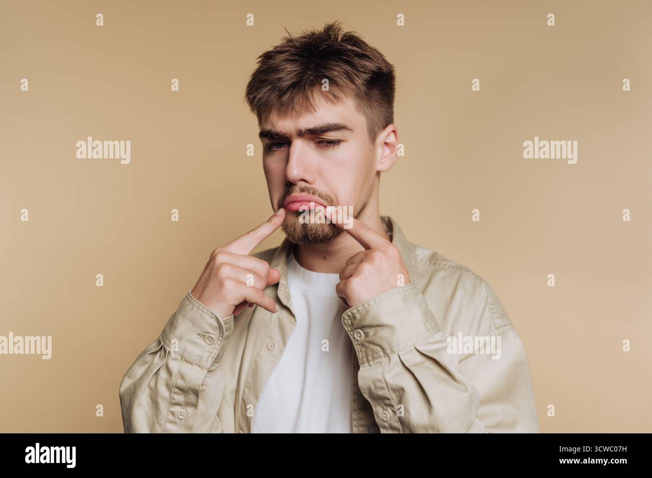 Das Porträt eines jungen Mannes zeigt einen traurigen Ausdruck, der die Verletzlichkeit betont, indem er auf seine Stirn auf beigefarbenem Hintergrund zeigt Stockfoto