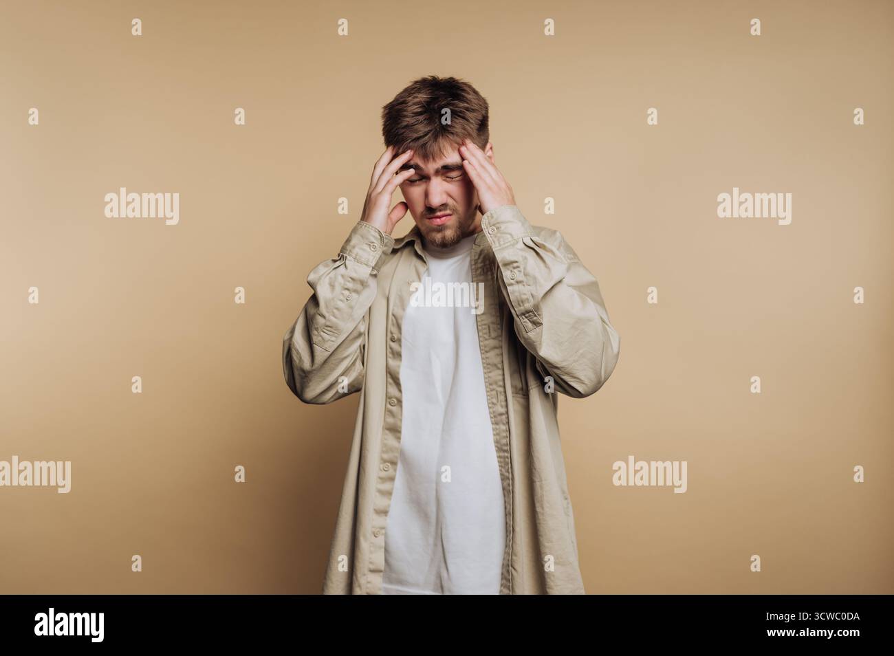Das Porträt eines jungen Mannes drückt Frustration aus, indem es seinen Kopf hält und einen emotionalen Moment auf beigefarbenem Hintergrund festhält Stockfoto
