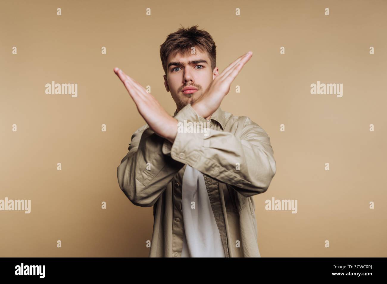 Porträt des jungen Mannes behauptet ein festes Nein mit gekreuzten Armen und vermittelt eine starke Botschaft der Ablehnung und Entschlossenheit auf beigefarbenem Hintergrund Stockfoto