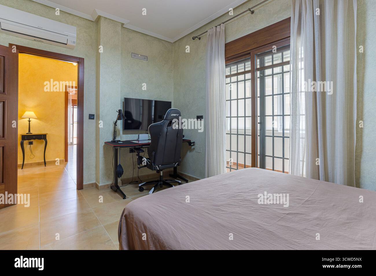 Ein stilvolles und funktionales Schlafzimmer mit ergonomischem Stuhl im Gaming-Stil und kompaktem Schreibtisch mit Monitor, Lampe und Zubehör. Das Zimmer inkl. Stockfoto