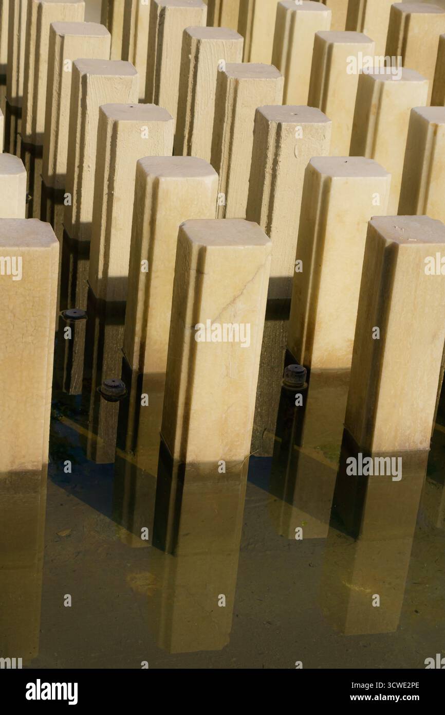 Serie rechteckiger Steinsäulen, die teilweise in Wasser getaucht sind und von Morgensonne beleuchtet werden. Die Reflexionen und sich wiederholenden Formen bilden ein Minimum Stockfoto