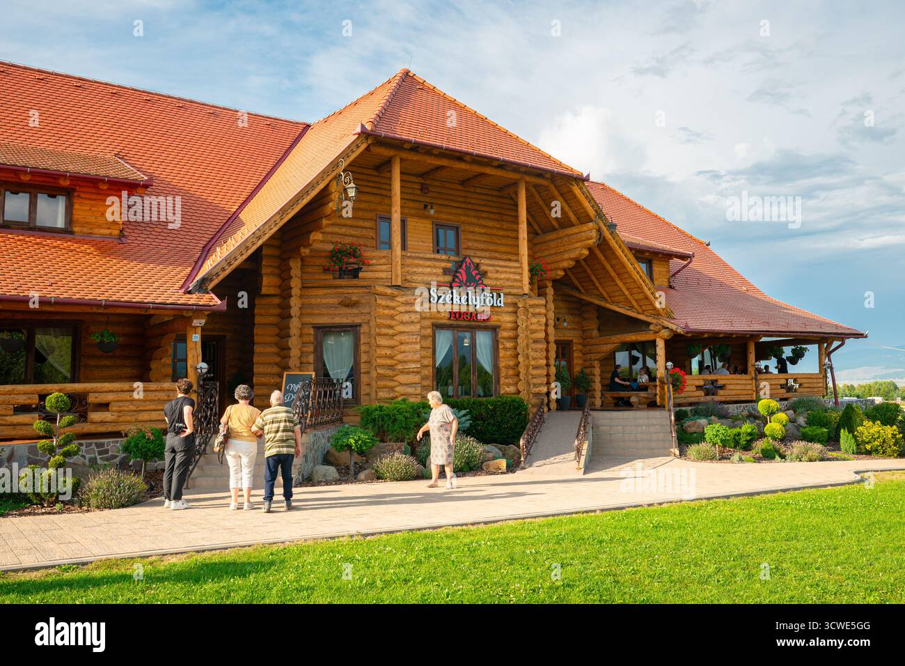 Gemütliches Blockhüttenrestaurant mit ankommenden Gästen, die einen sonnigen Tag auf dem Rasen in Borzont, Rumänien, genießen. Stockfoto