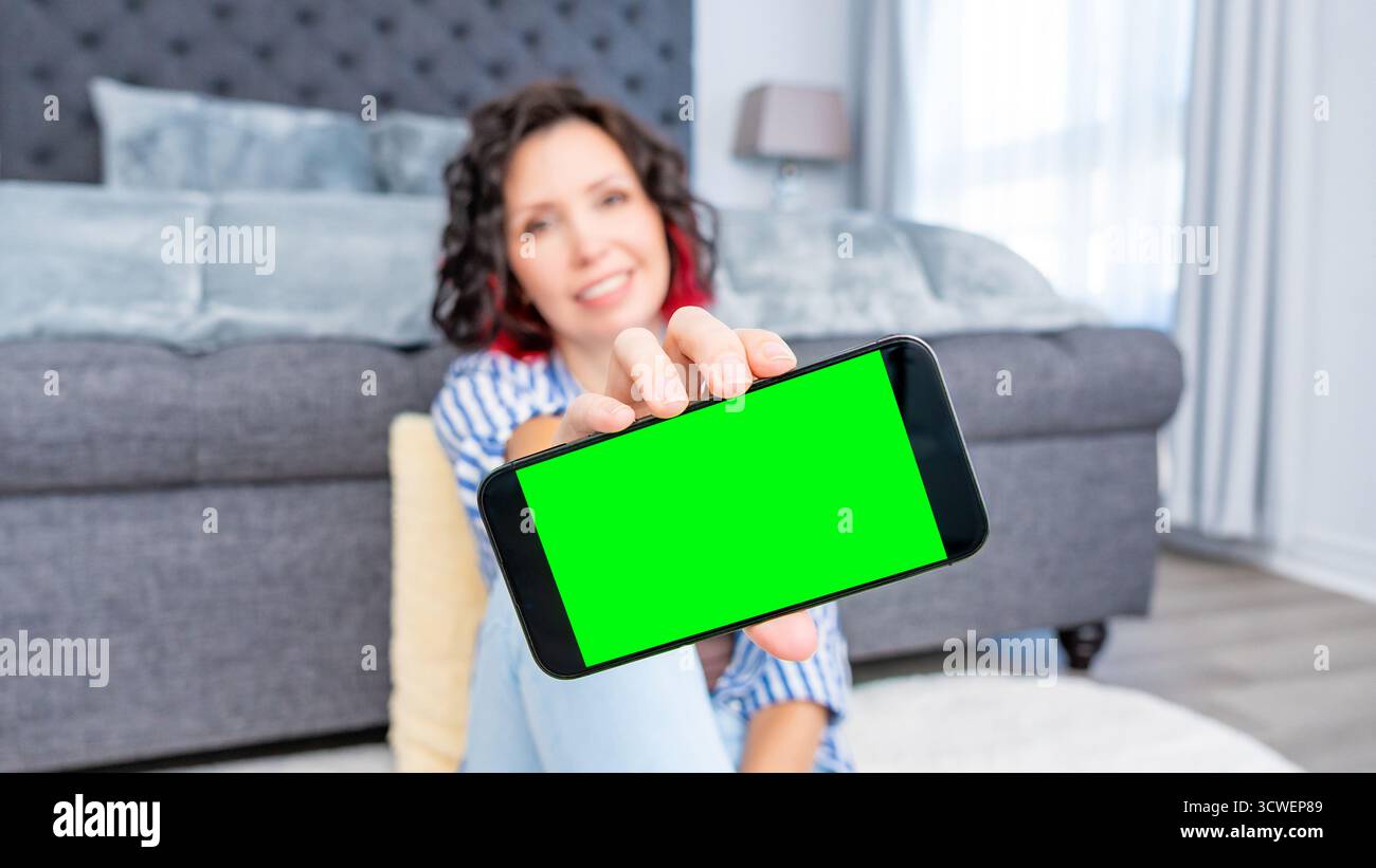 Frau in legeren Kleidern sitzt auf dem Boden, hält Smartphone-Mockup mit grünem Bildschirm in einem gemütlichen Wohnzimmer und hebt den zeitgenössischen Lebensstil hervor Stockfoto