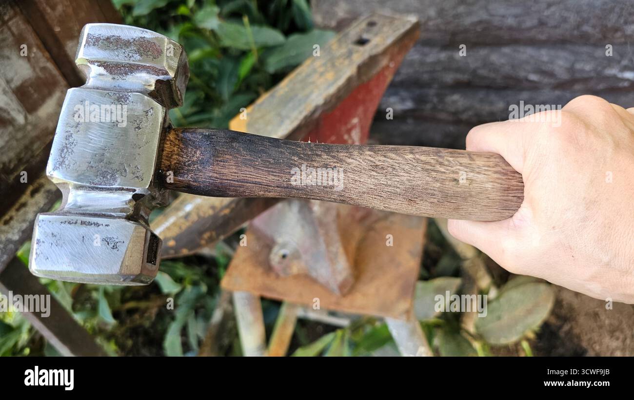Blick auf die Hand eines Mannes, der einen Schmiedehammer mit einem Amboss im Hintergrund hält. Stockfoto
