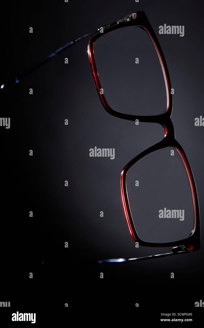 Studio-Stillleben von Brillen mit rotem Rahmen, der vertikal gegen dunklem verlaufendem Hintergrund hängt. Konzeptbild für modisches Accessoire und V Stockfoto