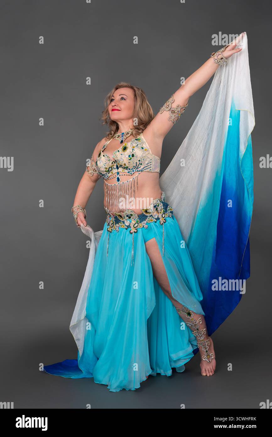 Lächelnde Bauchtänzerin mit Hand erhöht in Tanzbühnen-Outfit - BH-Oberteil und blauer langer Rock mit Schlitz und Pareo, der barfuß in voller Länge bewegt Stockfoto
