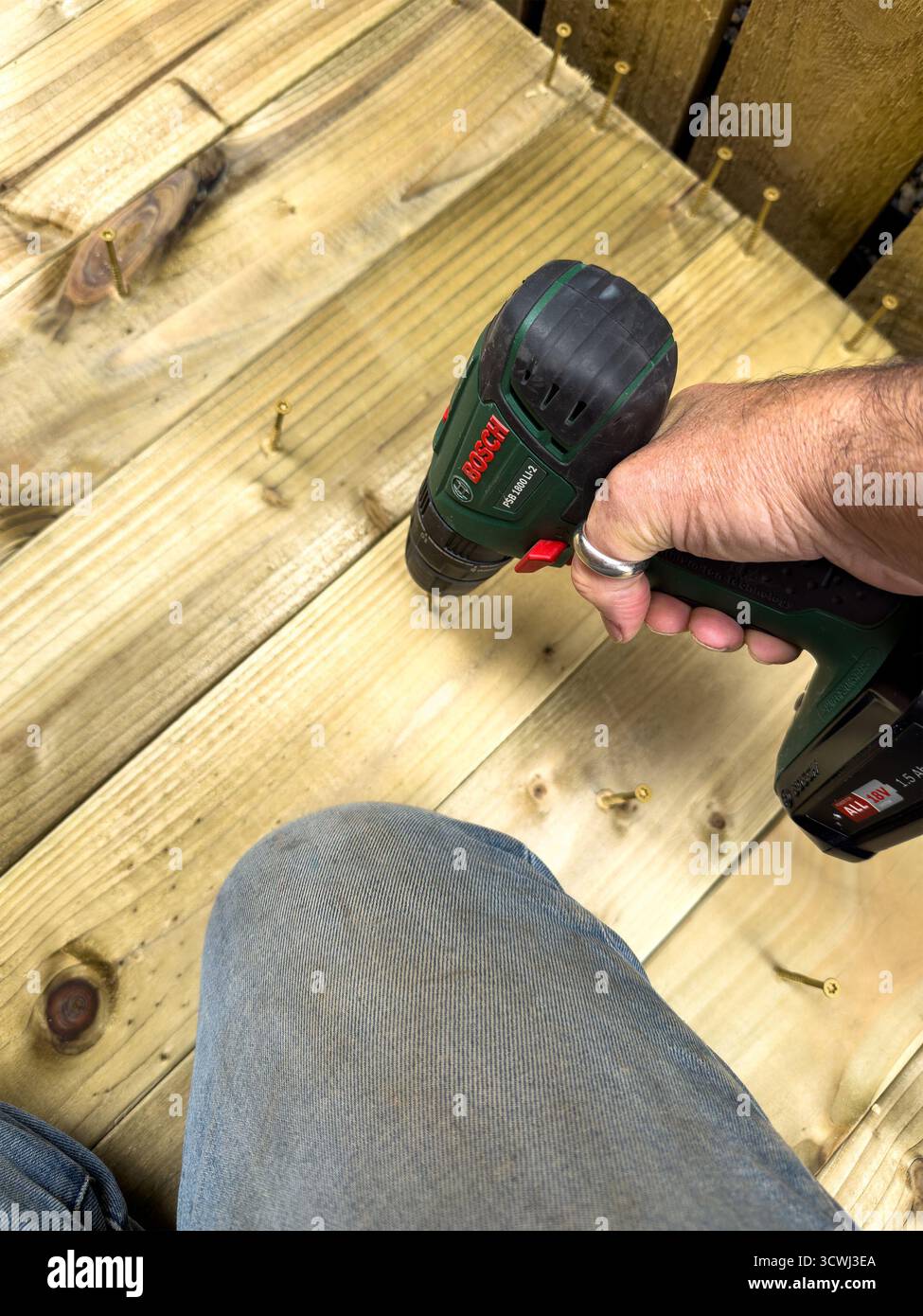 Person, die Holzdielen auf einem Deck mit einem Bosch Akku-Bohrer fixiert, wobei Hand und Werkzeug gegen die strukturierte Holzoberfläche gerichtet werden Stockfoto