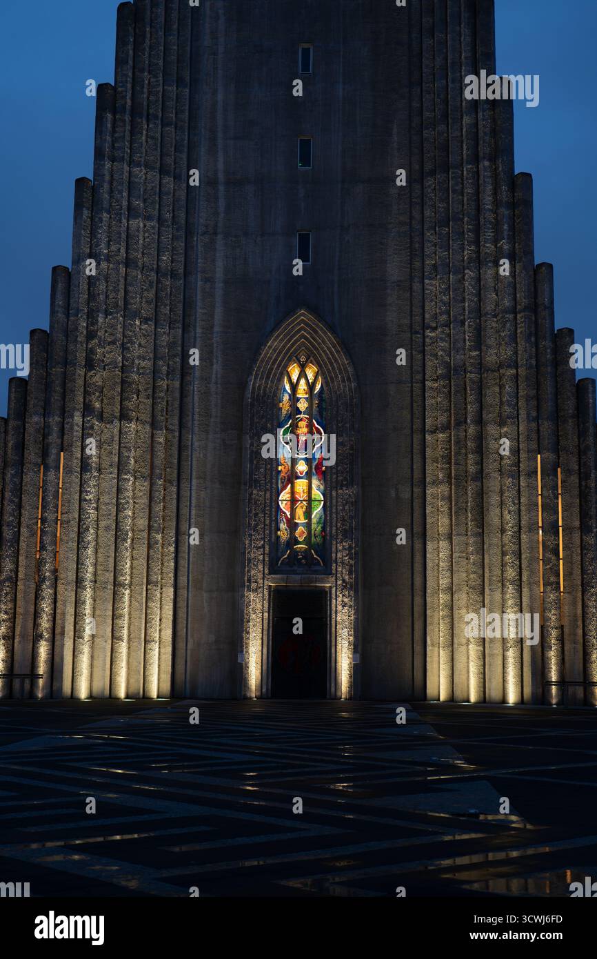 Hallgrimskirkja in Reykjavik, Island, verfügt über eine beleuchtete Fassade mit einem hohen Buntglasfenster, geometrischen Betonsäulen und einem gemusterten Vorderboden. Stockfoto
