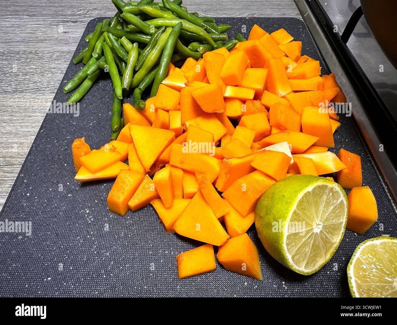 Gehackter Kürbis, frische grüne Bohnen und eine halbe Limette auf einem schwarzen Schneidebrett bereit zum Kochen Stockfoto