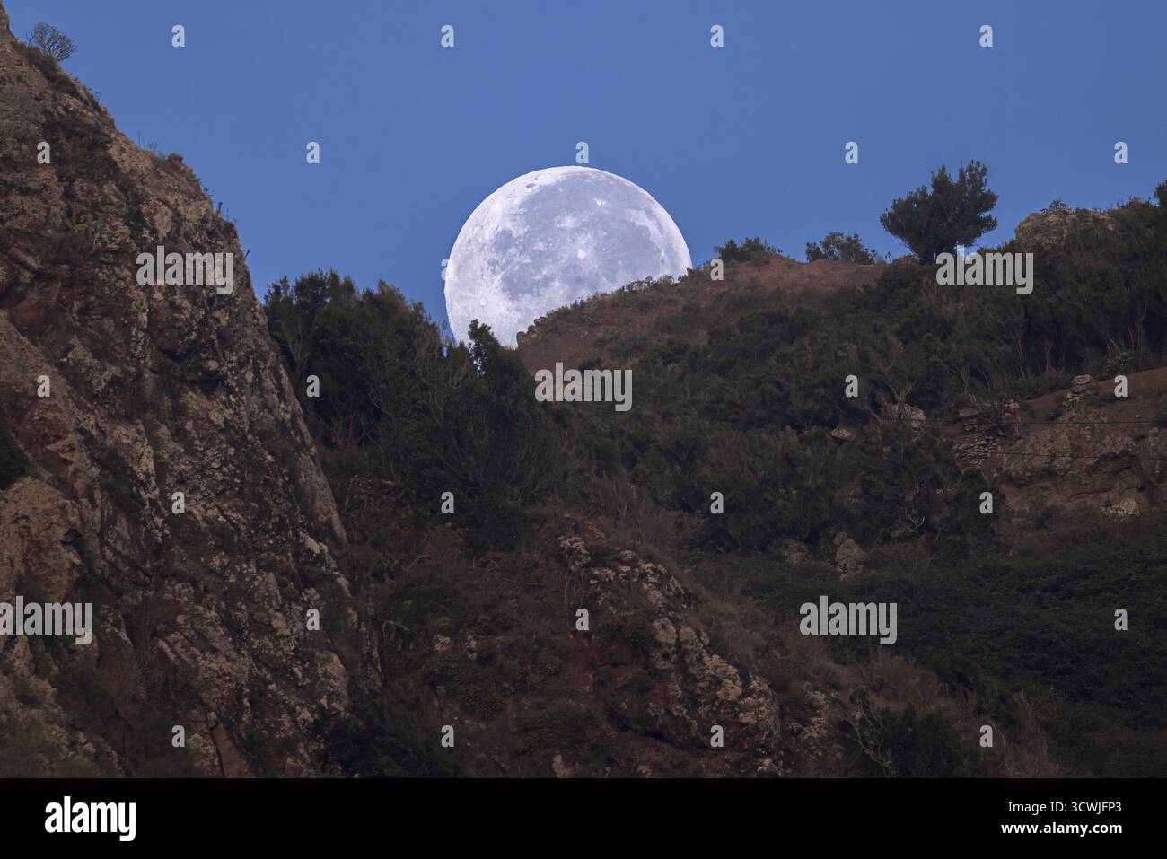 Ein atemberaubender Vollmond bestiegen die malerischen Gipfel der Anaga Mountains in der Dämmerung und schafft eine magische Atmosphäre auf Teneriffa, Spanien Stockfoto