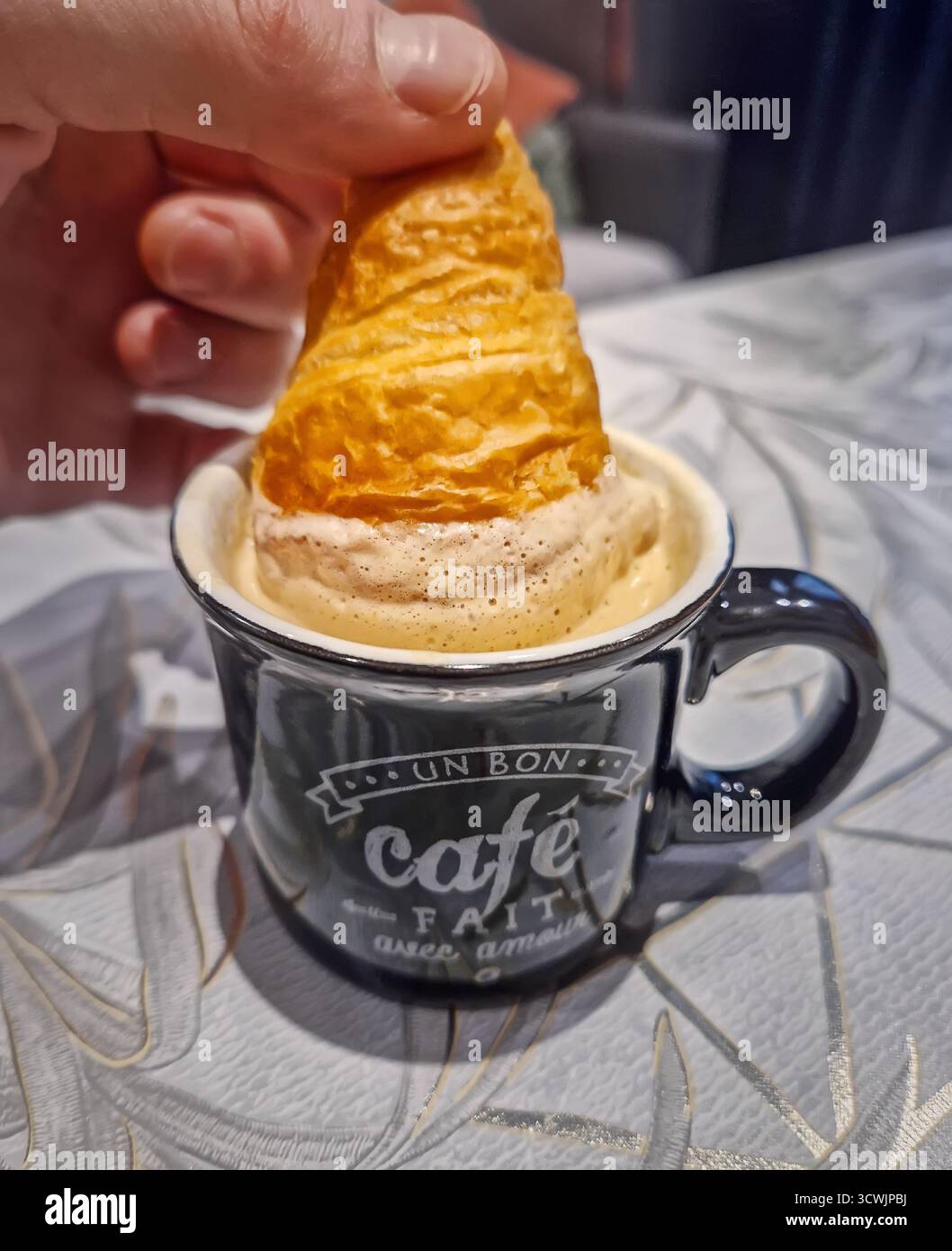 Eine Nahaufnahme taucht ein Mini-Croissant in eine schwarze Kaffeetasse, gefüllt mit einem aufschäumenden, karamellfarbenen Espresso-Getränk. Gutes Getränk mit reichhaltiger Creme Stockfoto