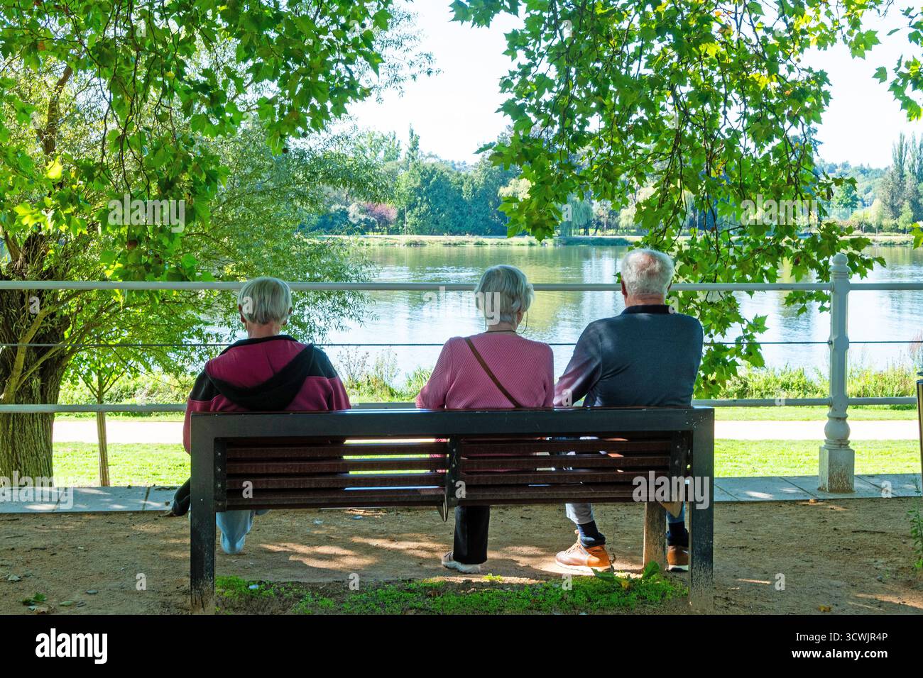 Vichy. Drei ältere Menschen sitzen auf einer Bank und blicken auf den Fluss Allier, während ein junges Paar Erinnerungen in der Nähe festhält. Allier. Auvergne . Frankreich Stockfoto