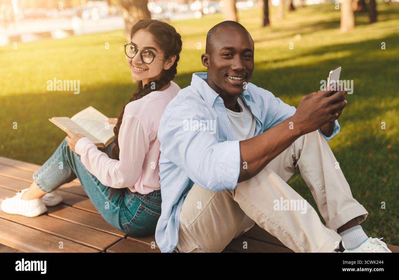 Glückliche, vielfältige Studenten, die sich mit dem Smartphone und dem Buch zurücklehnen und sich im Freien auf der Bank im Park ausruhen Stockfoto