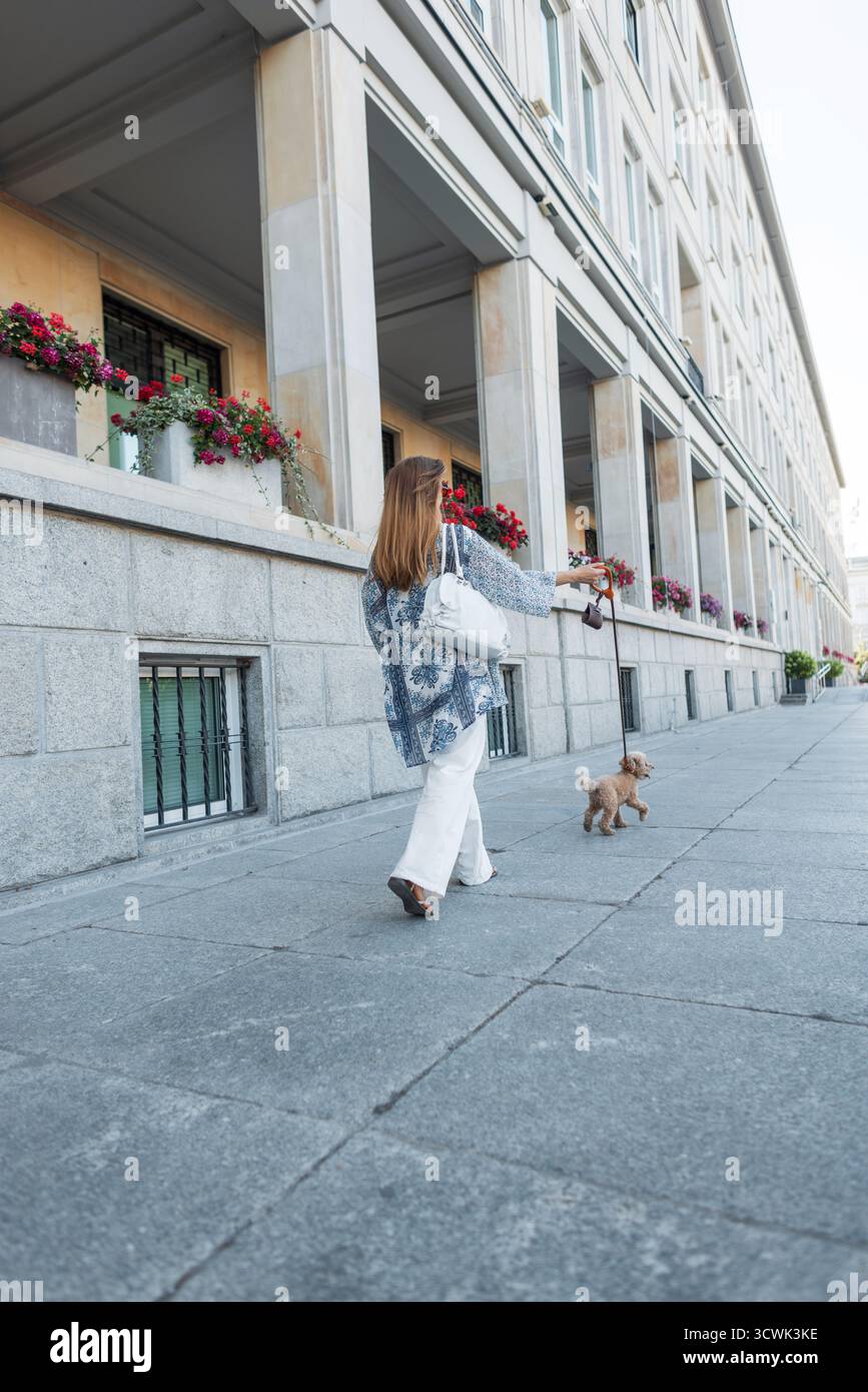 Eine Frau, die ihren kleinen Poodle an der Leine entlang Einer City Street führt. Rückansicht, Lässiges Sommeroutfit, Entspannter Urbaner Vibe. Pet Care, Daily Routine Und Moder Stockfoto