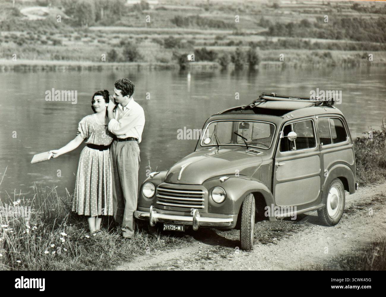 Romantische Kulisse für ein Foto, das ein junges italienisches Paar zeigt, das sich in der Nähe ihres Autos (ein FIAT Topolino Belvedere) an einem Fluss nahe der Stadt Mailand in den späten fünfziger Jahren umarmt Stockfoto