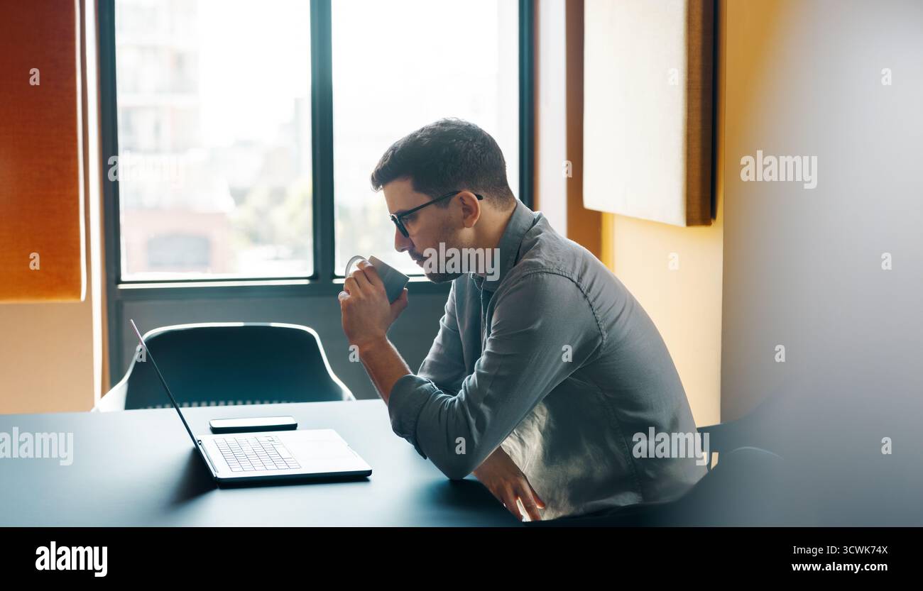 Ein Mann im Alter von 30 Jahren arbeitet an einem Laptop, während er in einer modernen Büroumgebung Kaffee trinkt. Der Laptop wird auf einem eleganten Tisch in der Nähe eines Fensters platziert Stockfoto