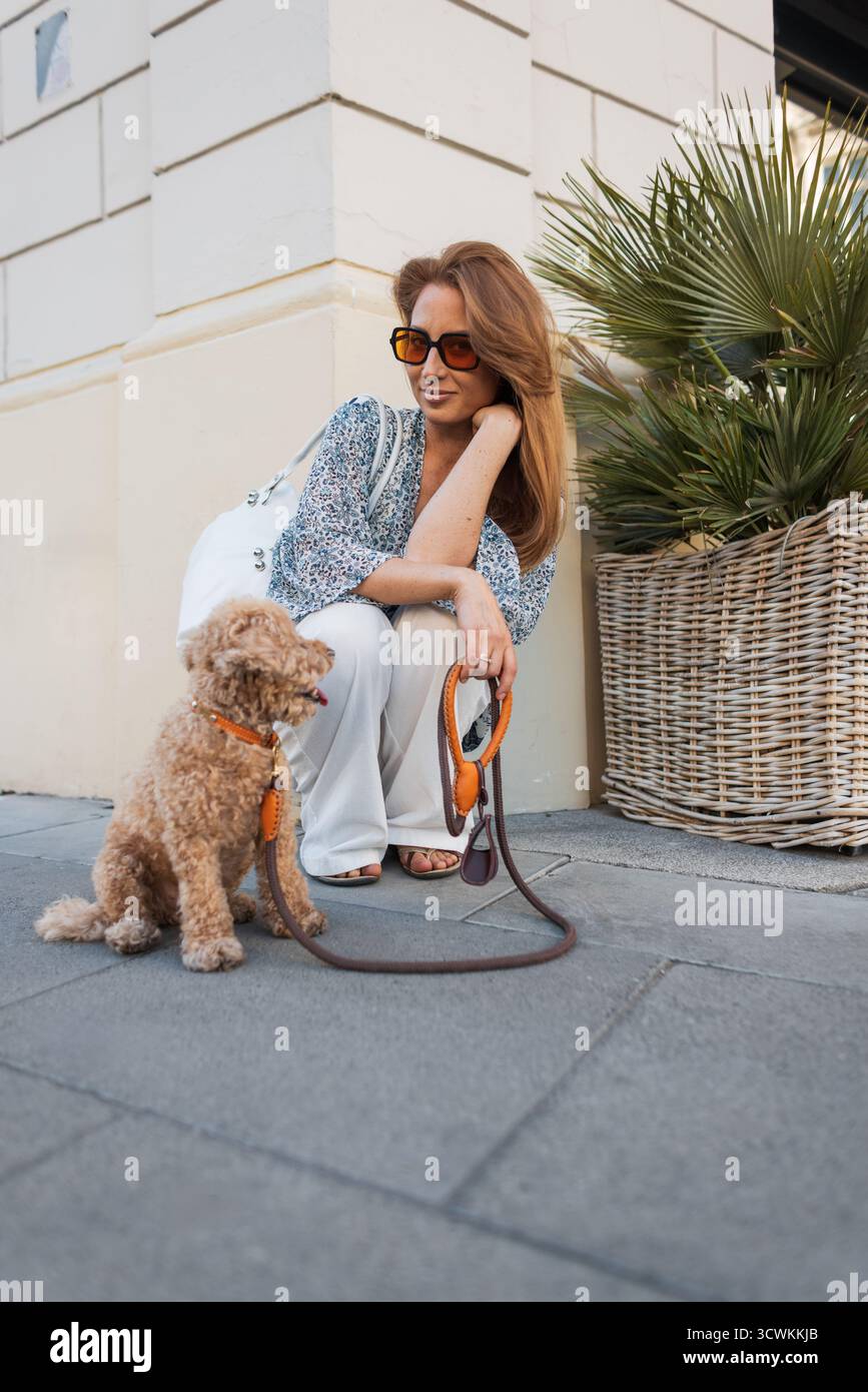 Eine Frau hockt sich auf Einem Bürgersteig der Stadt und gibt ihrem lockigen Hund Wasser aus Einer Portable Bowl. Warmes Wetter, urbaner Hintergrund und Ein zärtlicher Moment Stockfoto
