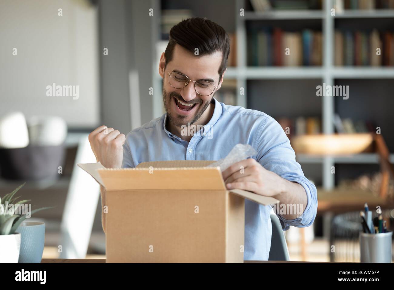 Ein glücklicher junger Mann, der das Postpaket am Schreibtisch öffnet Stockfoto