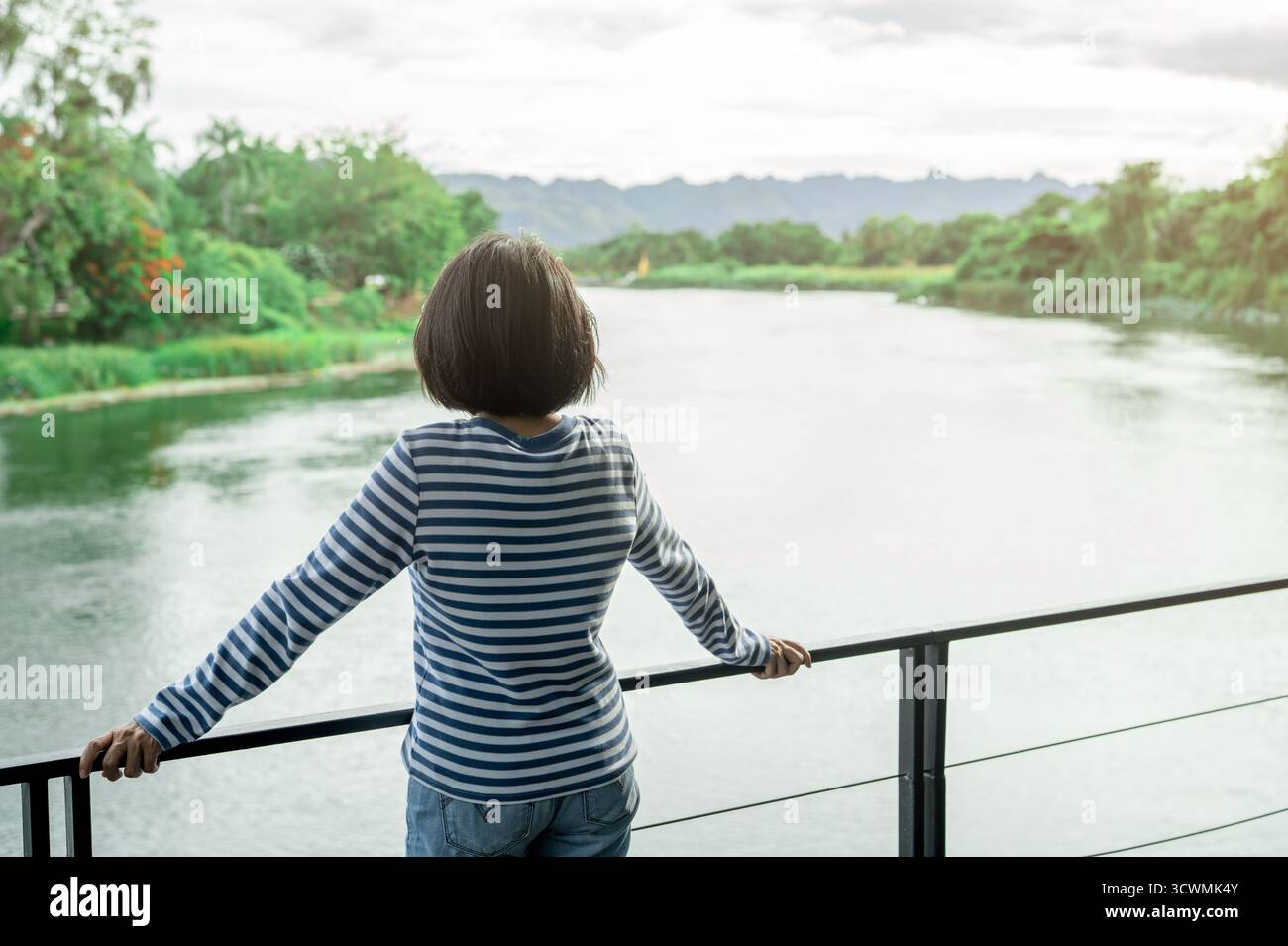 Frau, die auf einem Außenbalkon mit Blick auf den friedlichen Fluss und die Berge steht, Öko-Resort-Zuflucht für digitale Entgiftung, Naturverbundenheit und mentales Stockfoto