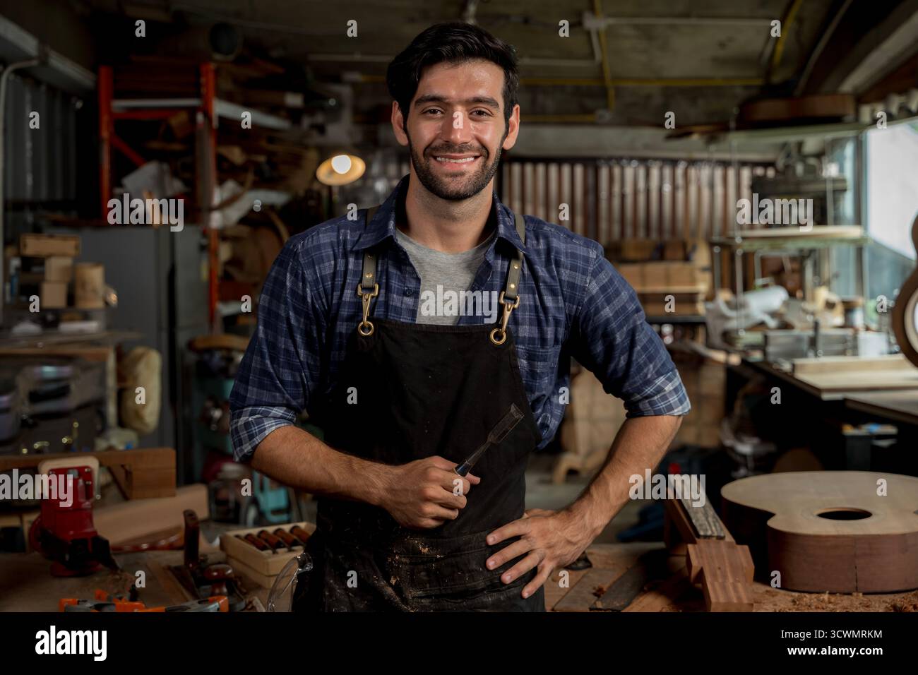 Selbstbewusster Zimmermann, der in der Werkstatt lächelt, während er einen Meißel hält, Sicherheitsschürze und lässiges Hemd trägt, was für Handwerkskunst, Kleinunternehmen oder Kleinunternehmen steht Stockfoto