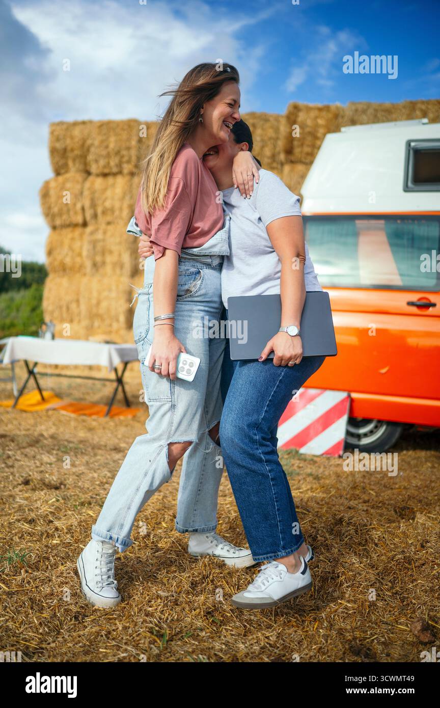 Zwei Frauen lachen und umarmen draußen neben einem orangen Wohnmobil und Heuballen. Stockfoto