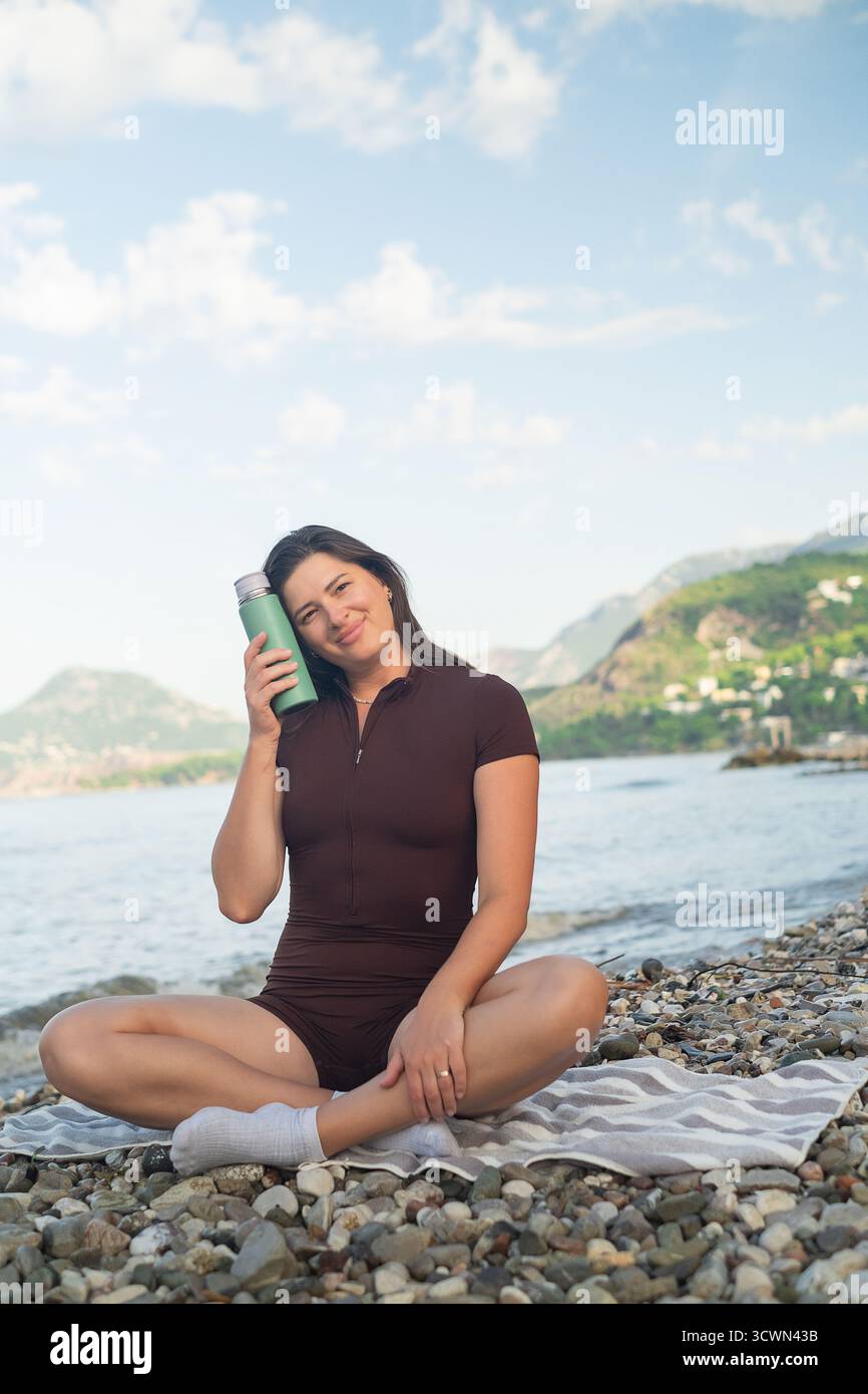 Gesundes Lifestyle-Konzept. Frau, die am Meer sitzt, sich ausruhen und nach dem morgendlichen Training mit Feuchtigkeit versorgt. Stockfoto