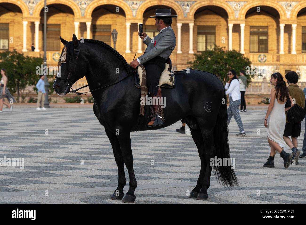 Sevilla, Spanien - 23. April: Ein Mann in traditioneller Kleidung, der auf einem schwarzen Pferd reitet und ein Smartphone hält, auf einem platz mit historischer Architektur im Hintergrund Stockfoto